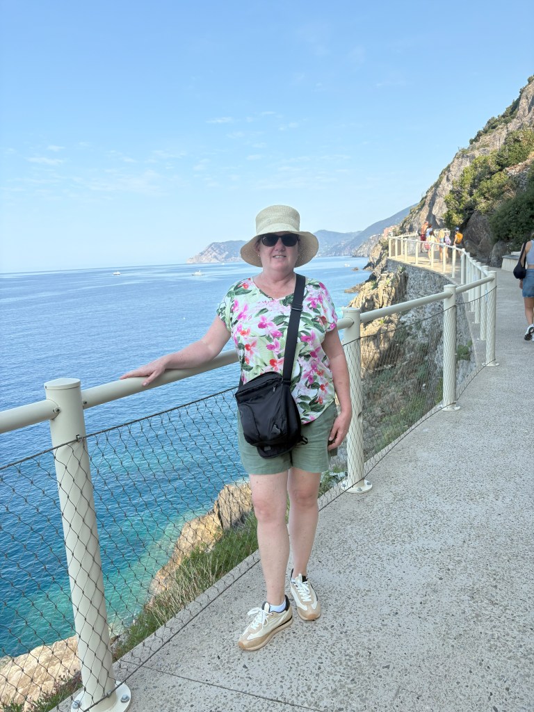 A woman in a floral shirt and shorts stands by a coastal pathway with a railing, overlooking the sea and mountains in the background.
