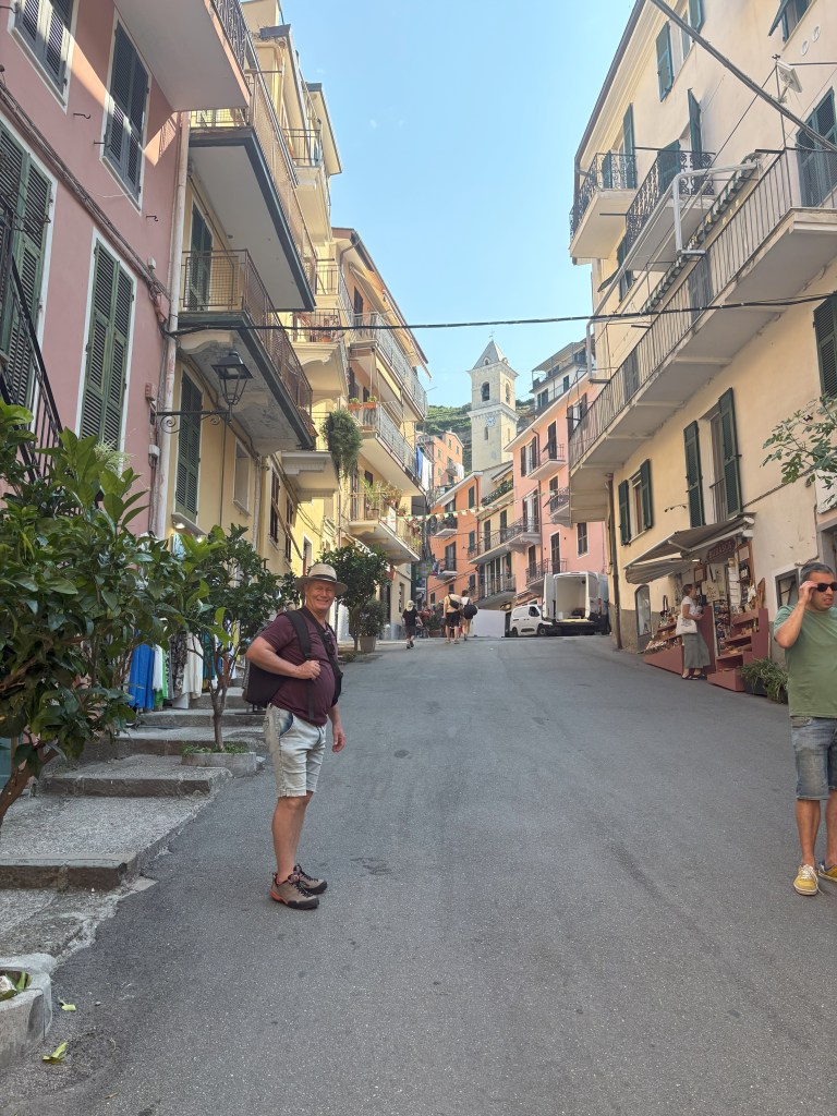 A narrow street lined with colorful buildings, featuring a man in a hat and shorts standing on the pavement, with additional pedestrians further up the hill and a church tower visible in the background.