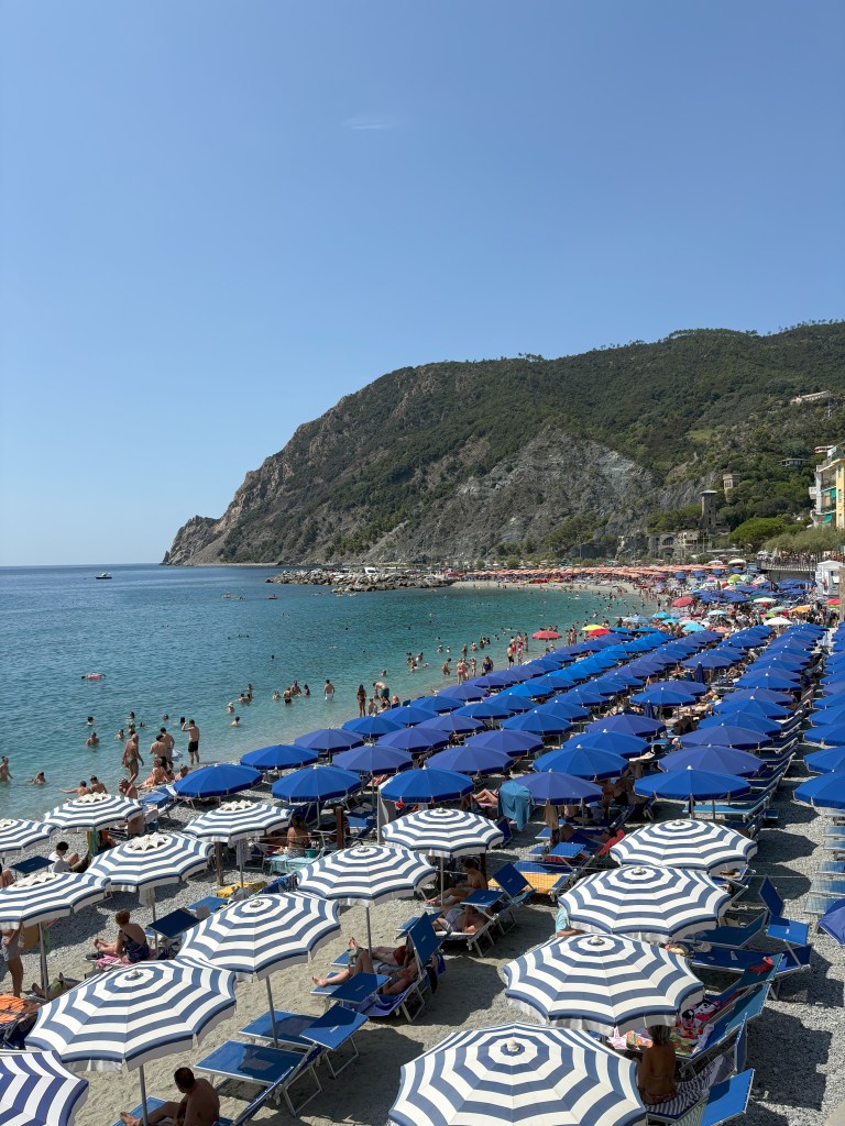 A busy beach scene featuring striped blue and white umbrellas and lounge chairs, with people swimming and relaxing by the water. A mountainous backdrop and clear blue sky complete the landscape.
