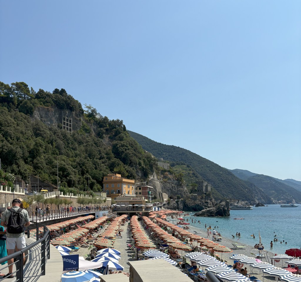 A beach scene featuring colorful umbrellas and sunbeds arranged in rows, with people enjoying the sun and swimming in the clear water. A rocky hill and buildings are visible in the background under a clear blue sky.