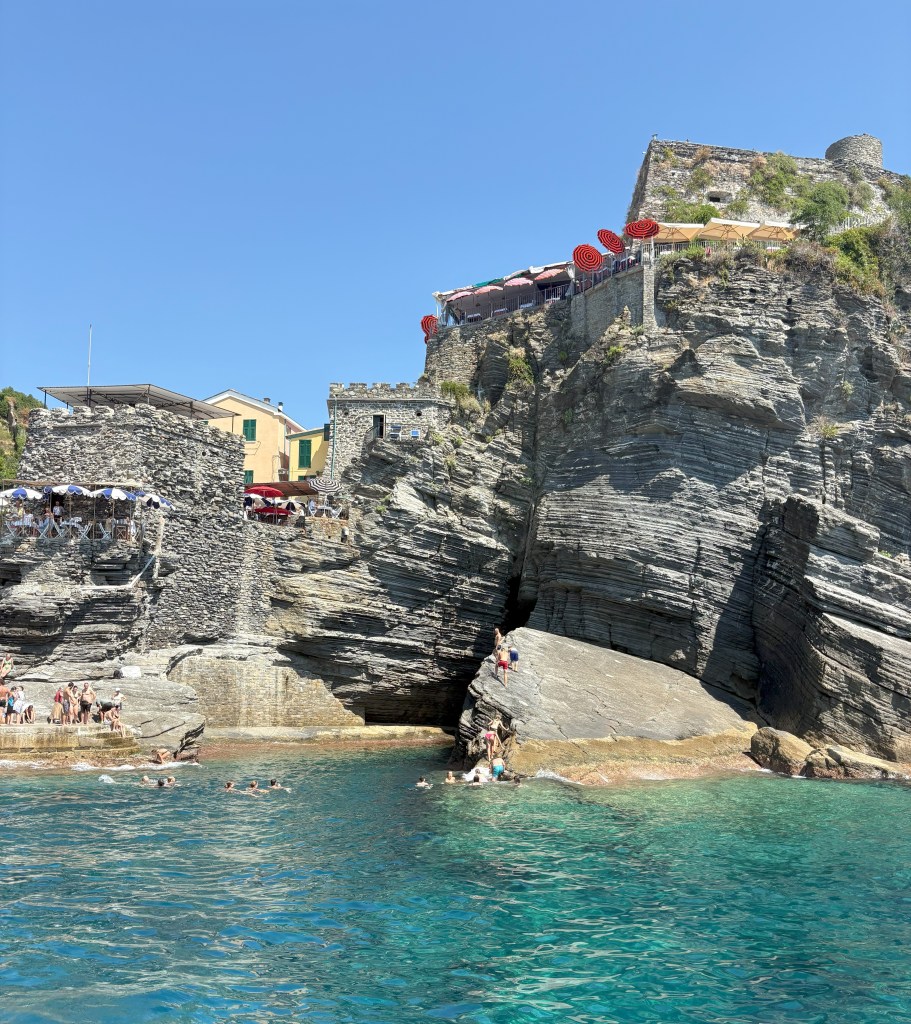 A rocky coastline with clear turquoise water, featuring historic stone buildings and a restaurant with umbrellas on a terrace.