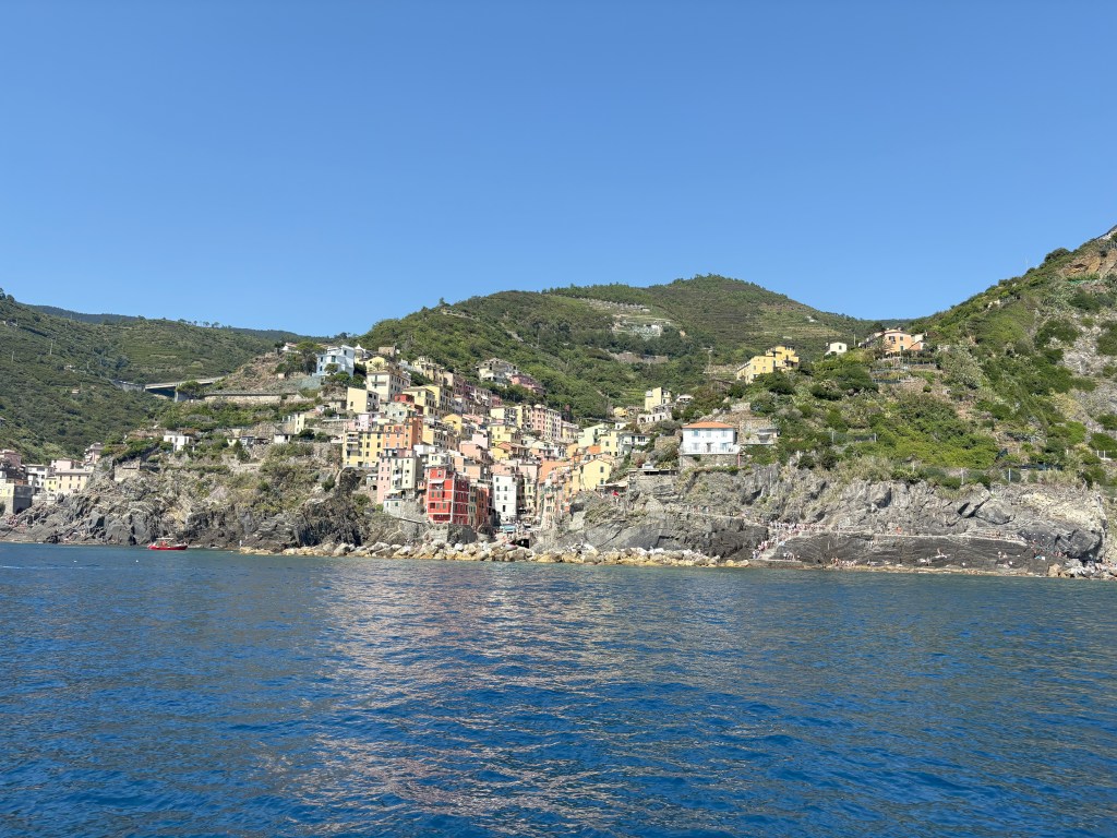 View of a coastal village with colorful buildings on a hillside, surrounded by green mountains and clear blue water.