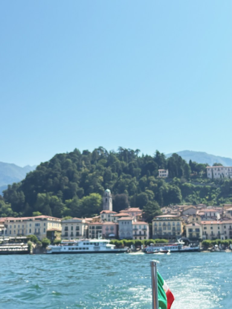 A view of a lakeside town with colorful buildings and lush green hills in the background, as seen from a boat on the water. A flag is visible in the foreground.