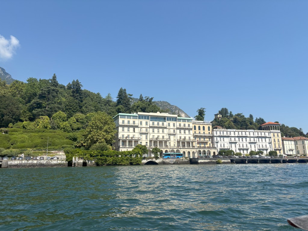Scenic view of a waterfront hotel along a lake, surrounded by lush greenery and mountains under a clear blue sky.