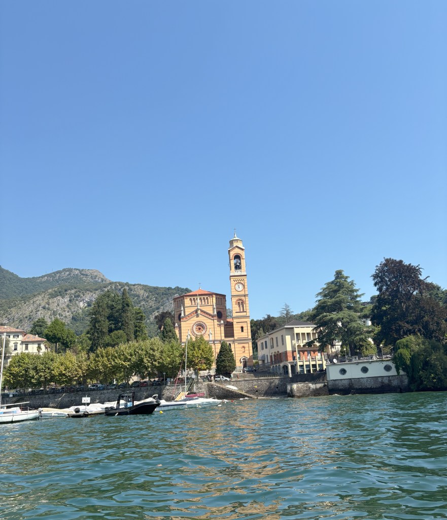 A picturesque view of a lakeside town with a clock tower and colorful buildings, surrounded by greenery and mountains under a clear blue sky.