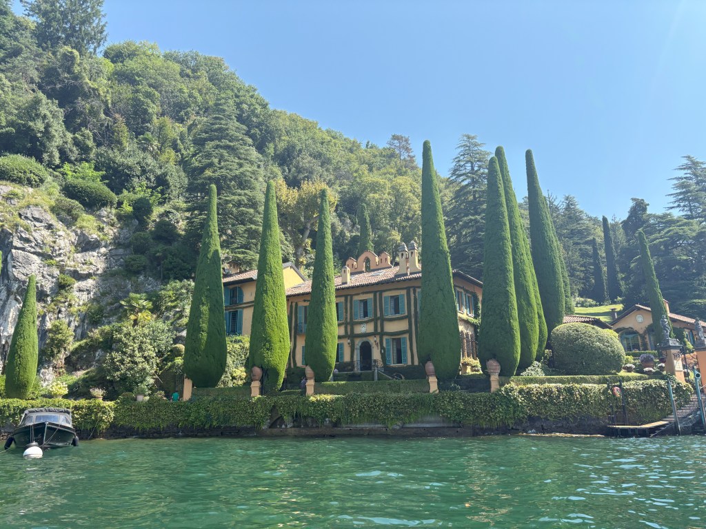 A picturesque lakefront villa surrounded by tall cypress trees and lush greenery, with clear blue water in the foreground.