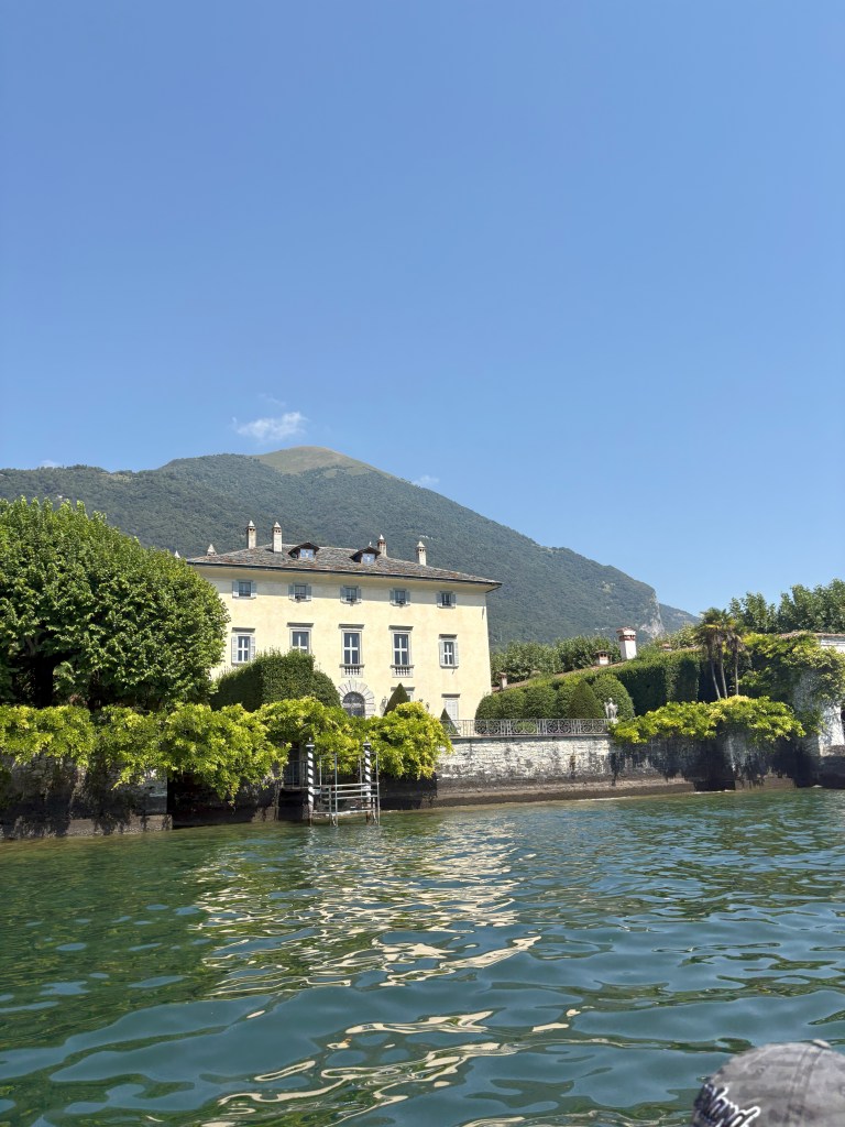 A scenic view of a lakeside villa surrounded by lush greenery, with a mountain in the background under a clear blue sky.