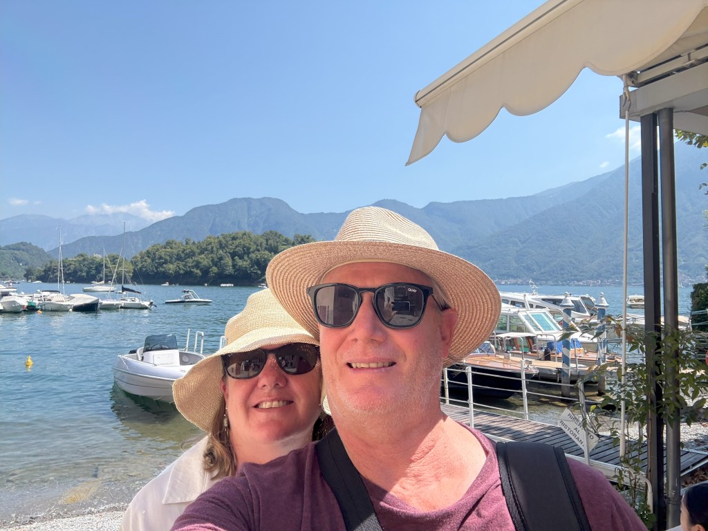 A smiling couple wearing hats and sunglasses, standing by the water with boats and mountains in the background on a sunny day.