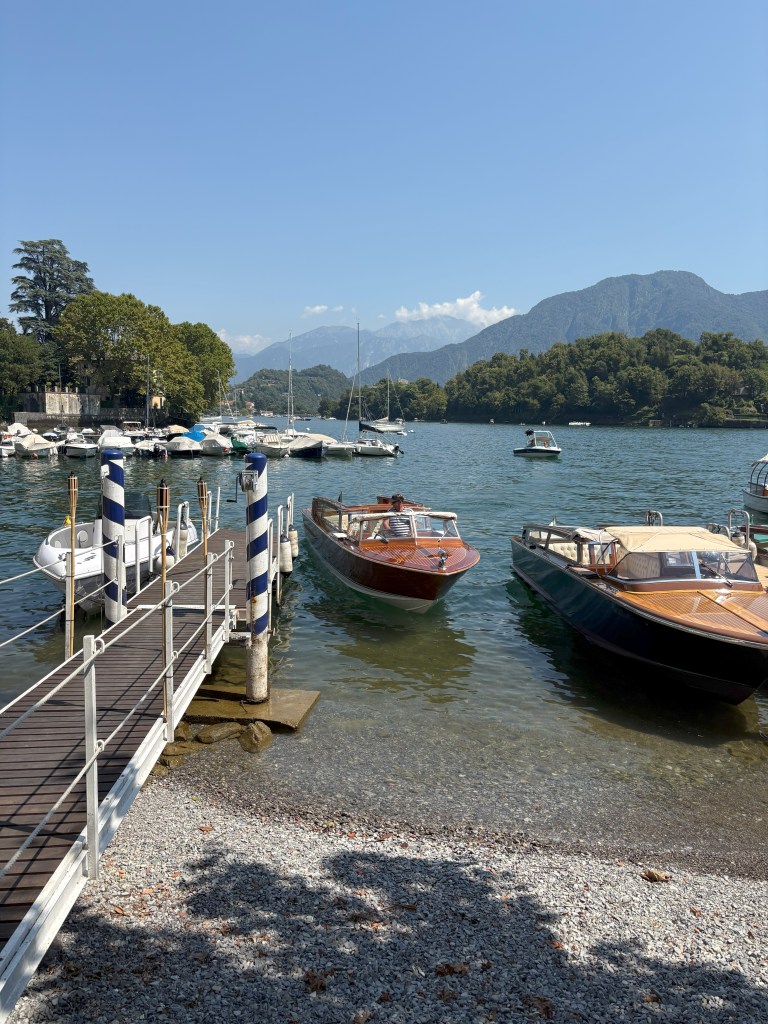 A scenic lakeside view with several boats docked near a wooden pier, surrounded by lush greenery and mountains in the background under a clear blue sky.