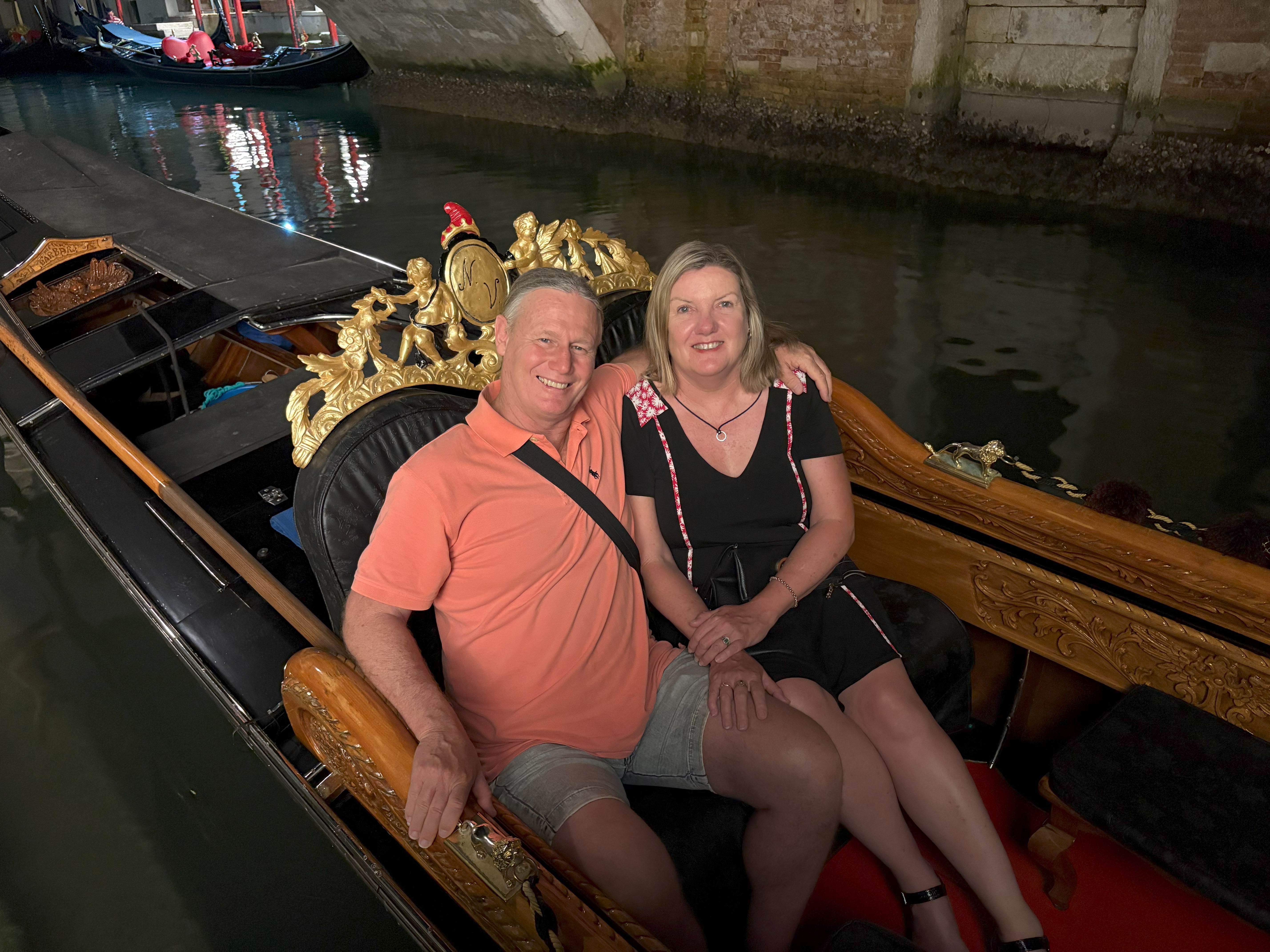Couple sitting on a decorated gondola in a canal at night, smiling at the camera