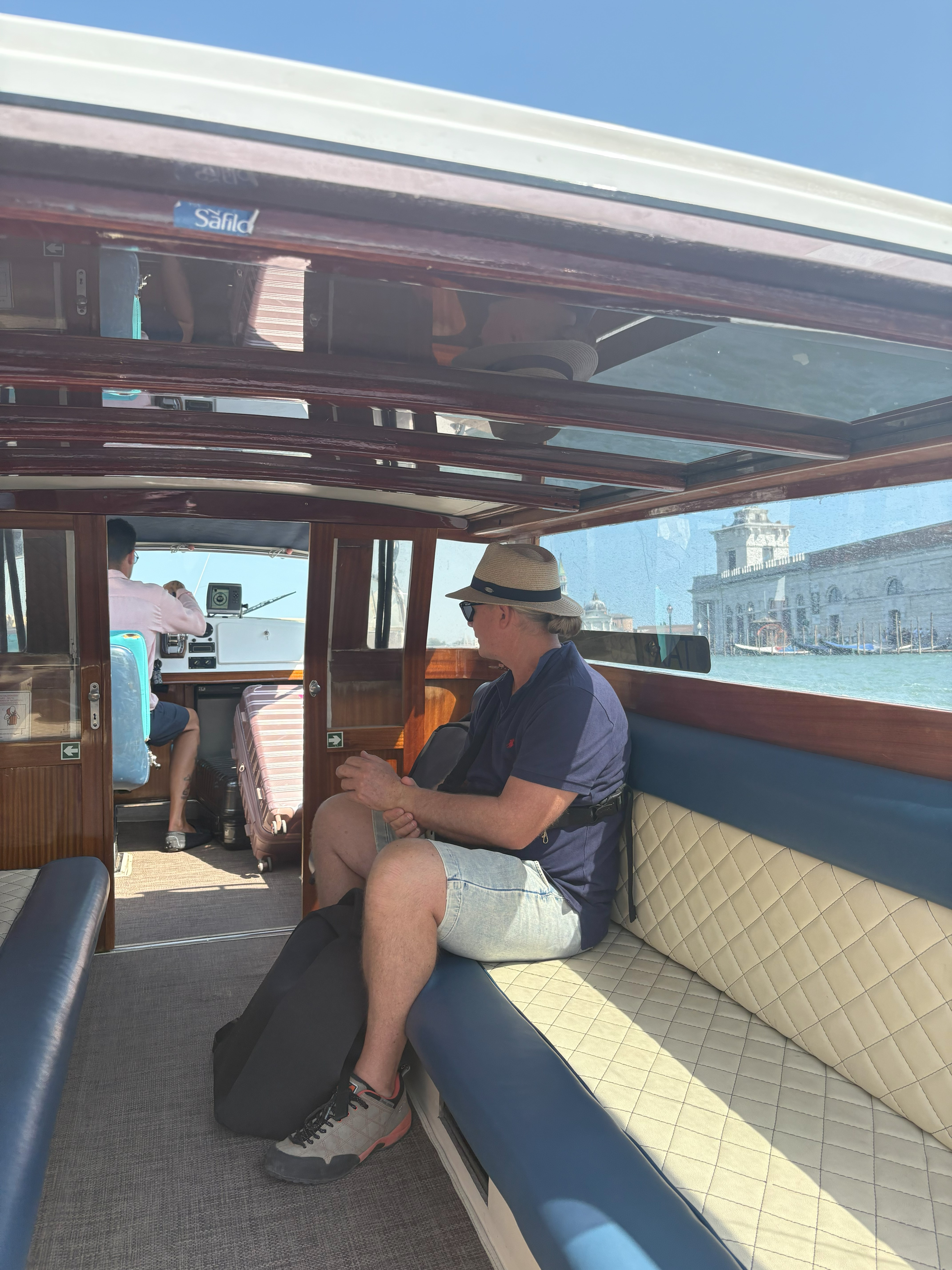 A man wearing a hat and sunglasses sitting inside a wooden boat with padded seats, looking out at the water and a historic building in the background